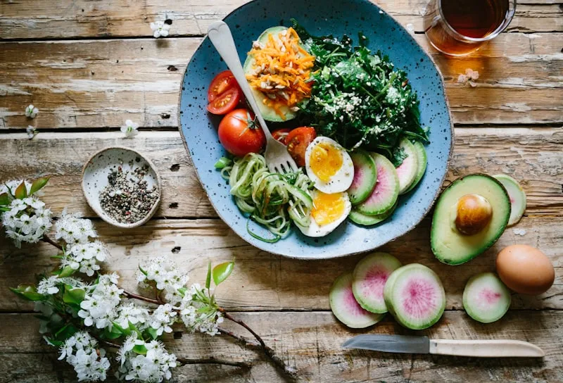 A blue plate with sliced avocado, hard-boiled egg, cherry tomatoes, greens, spiralized zucchini, and shredded carrots on a rustic wooden table with flowers and seasoning nearby.